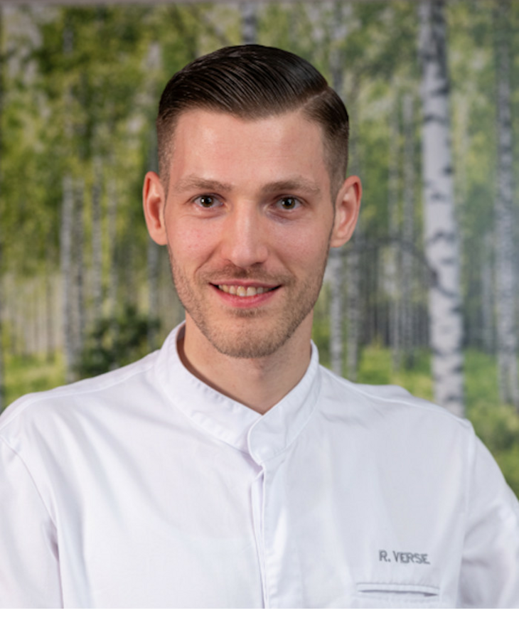 Chef René Verse | Tipken's restaurant by Nils Henkel on Sylt A chef in a white uniform smiles in front of a background with a birch forest motif.