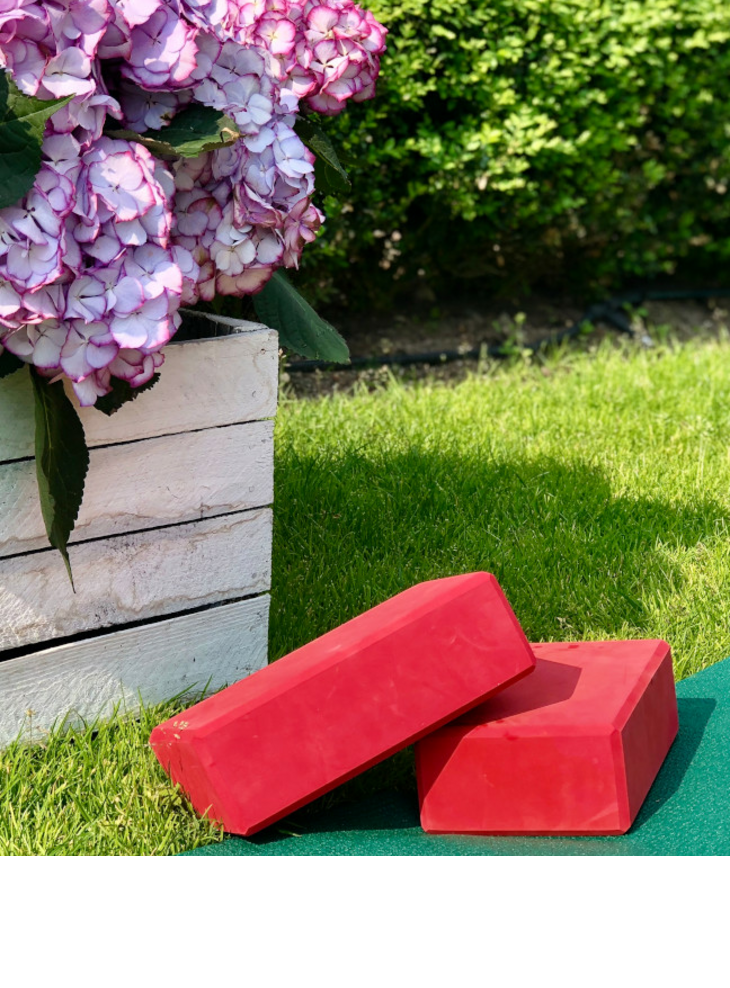 A white wooden box with pink and purple hydrangeas stands on a green lawn. Next to it, two red yoga or fitness blocks lie on the grass.