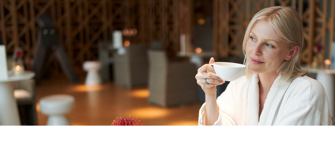 Eine Frau im Bademantel sitzt im Spa Café und Trinkt aus einer weißen Tasse