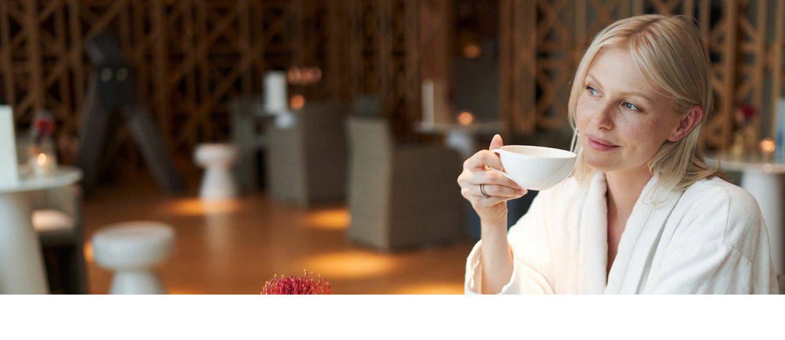 A woman in a bathrobe sits in the spa café and drinks from a white cup