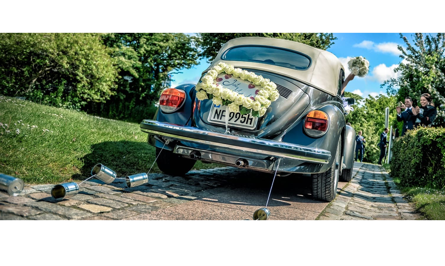 VW Käfer des Brautpaares mit "just married" Dekoration vor der Kirche St. Severin in Keitum Wedding car with flower wreath and cans at the rear, driving on a paved road, surrounded by green landscape.